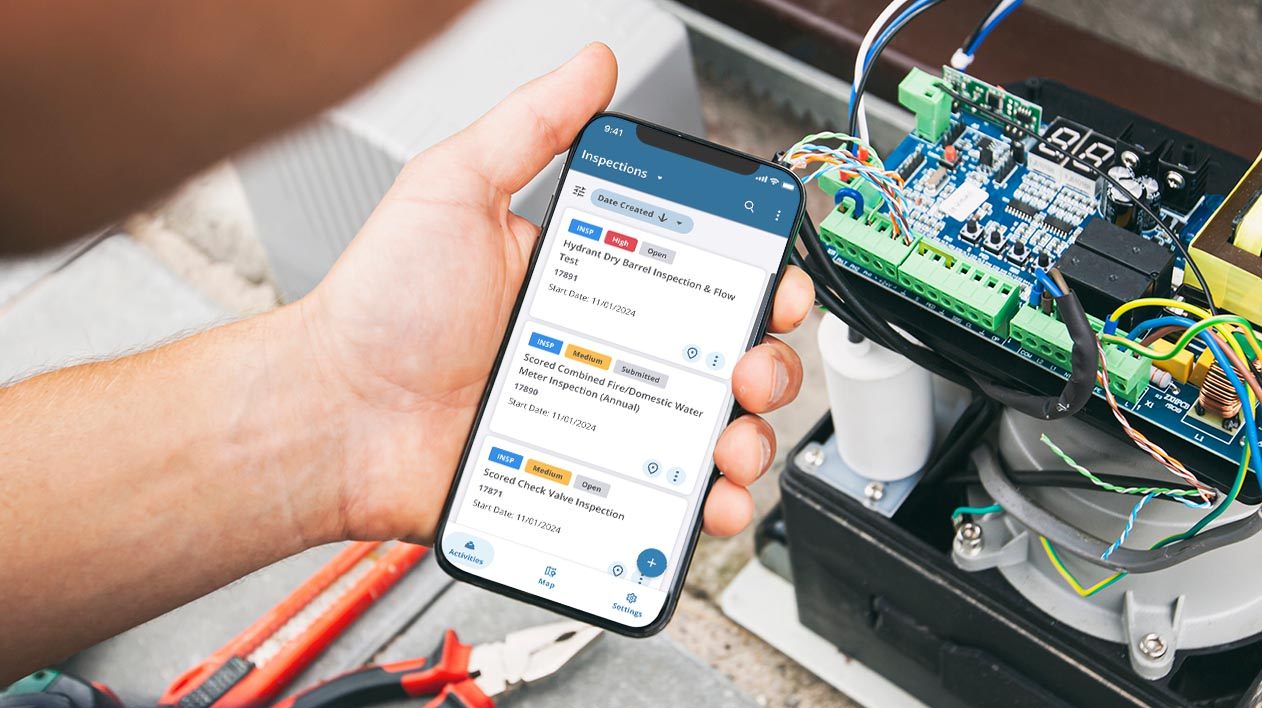 A technician holds a smartphone displaying a list of inspections, while working on an open electrical box with visible wires and a circuit board in the background.