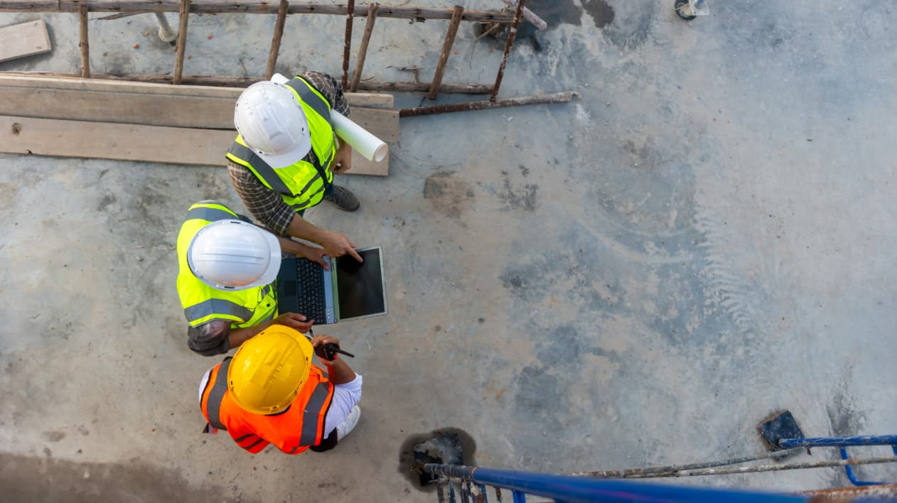 High-angle view of three construction professionals in safety gear reviewing a laptop and plans on site.