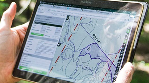A person holds a tablet displaying a digital map with topographical lines and paths.