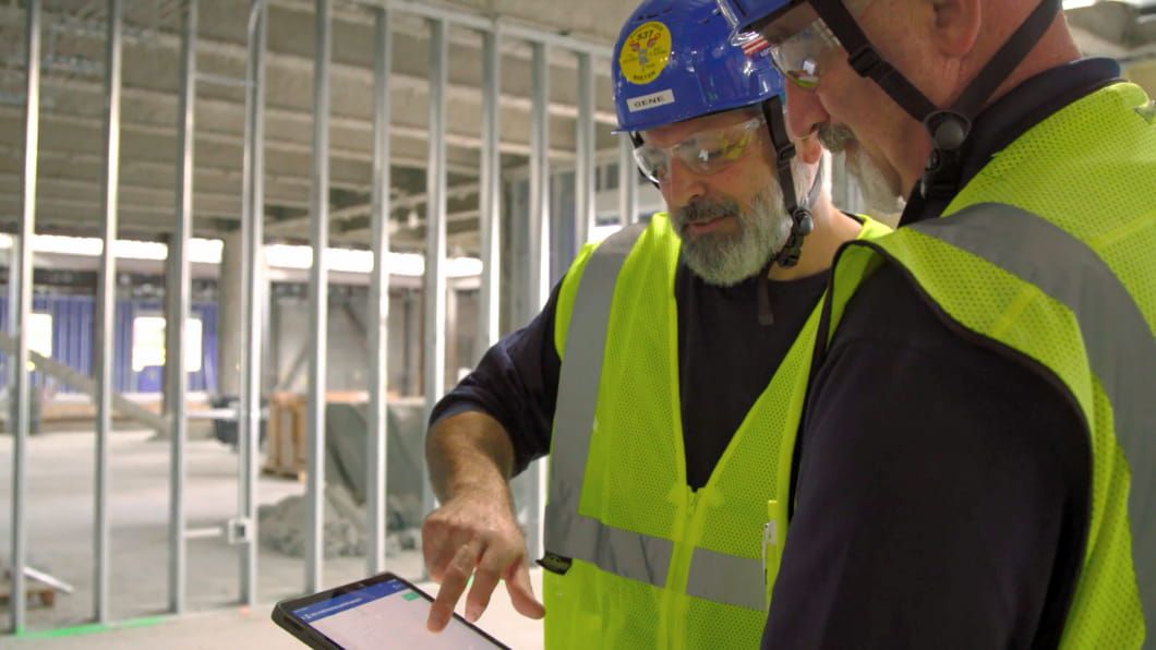 Two F+F Mechanical workers in safety gear review plans on a digital tablet at an indoor building site.