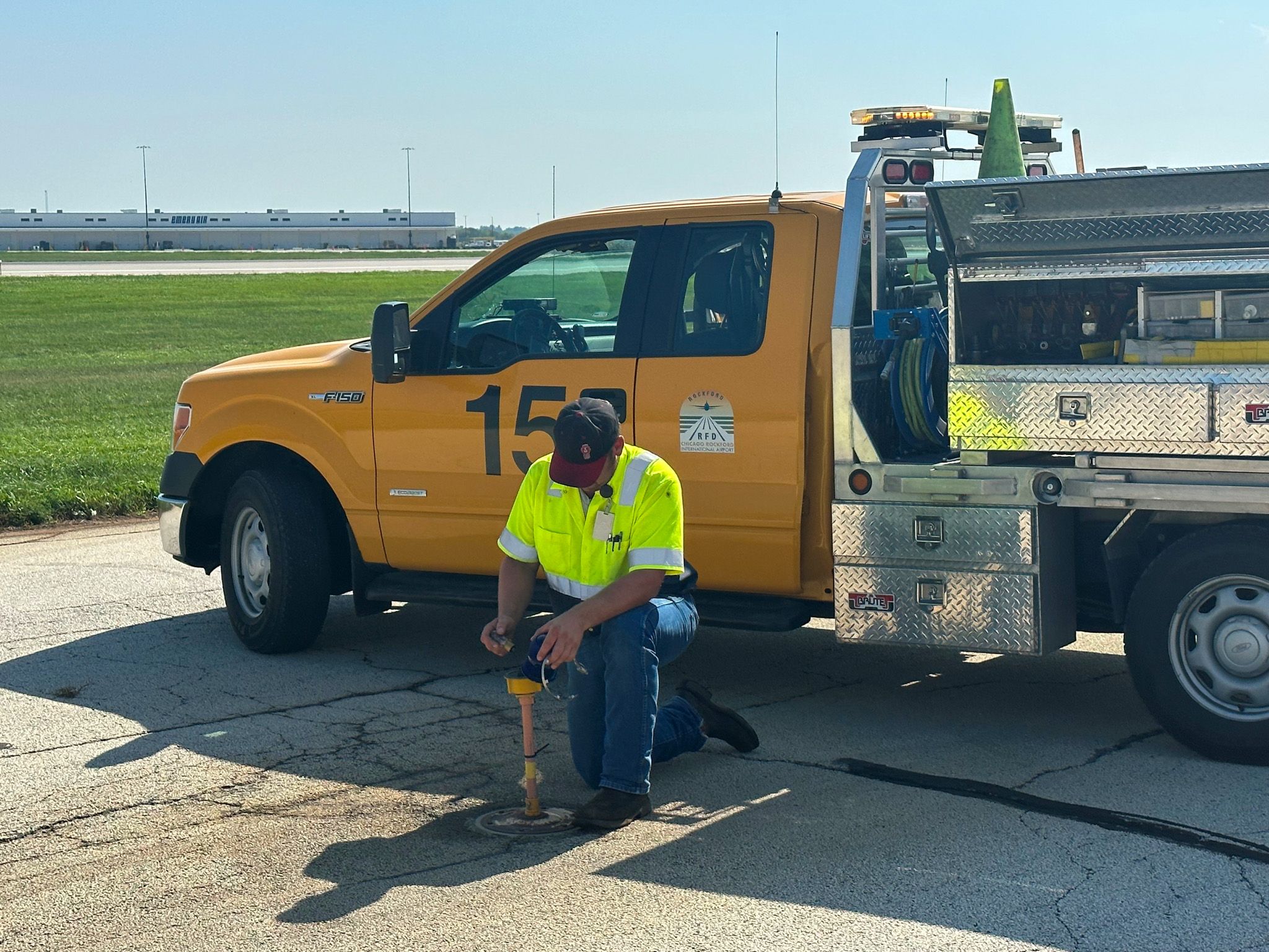 Maintenance worker in hi-vis vest uses jackhammer on airfield next to a yellow truck.
