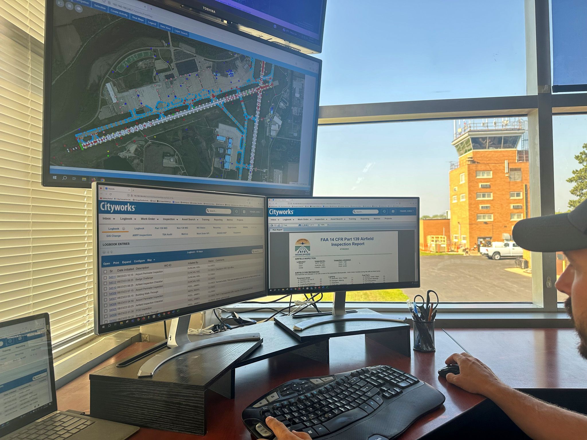 Person using multiple computer monitors displaying airport maps and reports, with an airport control tower visible outside.